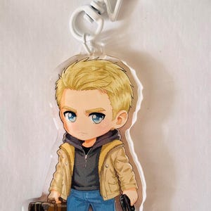 May include: An acrylic keychain featuring a cartoon character with blonde hair, blue eyes, and a serious expression. The character wears a tan jacket over a dark hoodie, blue jeans, and light shoes, holding a brown suitcase and a black handgun.