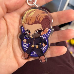 May include: A clear acrylic keychain featuring a cartoon character with blonde hair, a black mask, and a black uniform. The character is set against a blue and white badge. The keychain has a white star-shaped clasp.