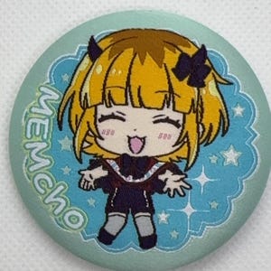May include: A round button with a cartoon illustration of a character with yellow hair, black horns, and a dark outfit. The button has a light blue background with white stars and the word "MEMCHO" in green.