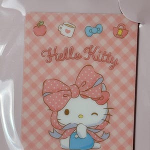 May include: A rectangular Hello Kitty card mirror in a pink and white gingham pattern. The mirror features a graphic of Hello Kitty wearing a large pink polka-dot bow and a blue outfit. Small icons of apples, mugs, and bows surround the character.