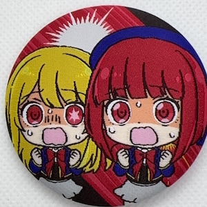 Peut inclure: Un écusson rond brodé représentant deux personnages de style anime avec des expressions surprises. L'un a les cheveux jaunes, l'autre rouges. Les deux portent des tenues bleues et rouges, sur fond rouge et noir.