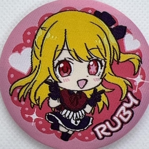 May include: A round, pink button featuring a cartoon anime character with yellow hair and red eyes. The character is wearing a dark red and black outfit. The word "RUBY" is written in white on the button.