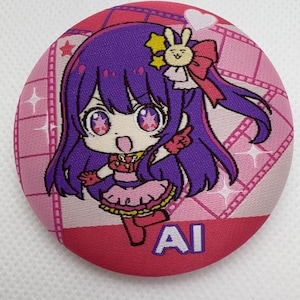 以下が含まれることがあります： 紫色の髪、大きな目、ピンクのドレスを着たアニメの漫画のキャラクターが描かれた、丸いピンク色のボタン。ボタンの下部には「AI」の文字があり、周囲にはフィルムストリップのデザインがあります。