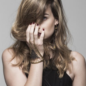 Puede incluir: Una mujer con el pelo largo y ondulado castaño lleva un top negro sin mangas con un patrón geométrico. Lleva una pulsera de oro y negro en la muñeca izquierda y pendientes de oro.