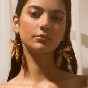May include: A woman with long dark hair wearing large gold earrings with delicate white flower accents. The earrings are made of dried leaves and flowers.