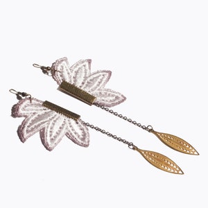 Peut inclure: Une paire de longues boucles d'oreilles délicates avec un motif floral. Les boucles d'oreilles présentent une fleur en dentelle blanche avec un centre rose, une barre dorée et une chaîne avec un pendentif en forme de feuille dorée.