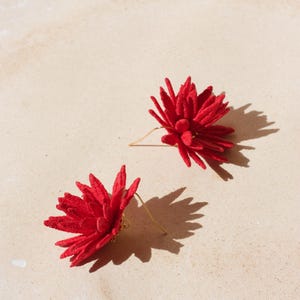 Puede incluir: Un par de pendientes rojos con forma de flor. Cada pendiente presenta un dise&ntilde;o detallado y texturizado con un gancho dorado. Los pendientes est&aacute;n colocados sobre un fondo neutro y claro, con sombras.