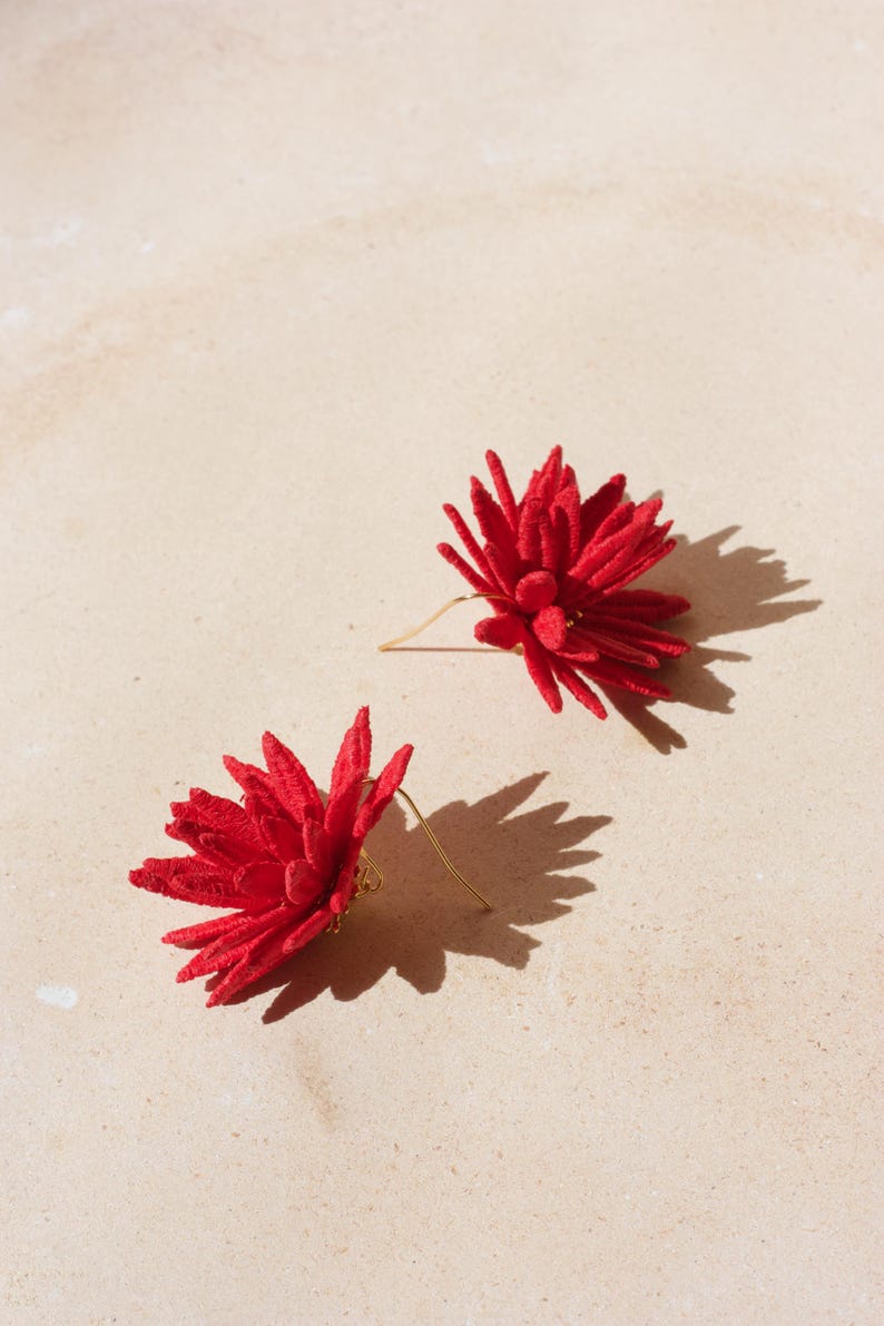 Puede incluir: Un par de pendientes rojos con forma de flor. Cada pendiente presenta un dise&ntilde;o detallado y texturizado con un gancho dorado. Los pendientes est&aacute;n colocados sobre un fondo neutro y claro, con sombras.