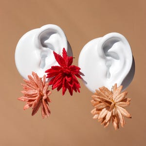 Puede incluir: Dos esculturas de orejas de cer&aacute;mica blanca adornadas con elementos florales vibrantes. Una oreja presenta una flor roja, la otra una flor melocot&oacute;n y marr&oacute;n claro. El fondo es de color beige liso.