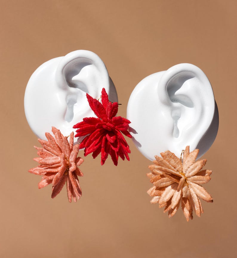 Puede incluir: Dos esculturas de orejas de cer&aacute;mica blanca adornadas con elementos florales vibrantes. Una oreja presenta una flor roja, la otra una flor melocot&oacute;n y marr&oacute;n claro. El fondo es de color beige liso.