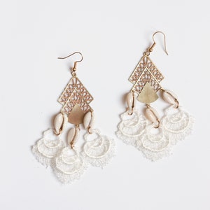 May include: A pair of gold earrings with white shell accents and delicate white lace fringe. The earrings feature a geometric design with intricate cutouts.