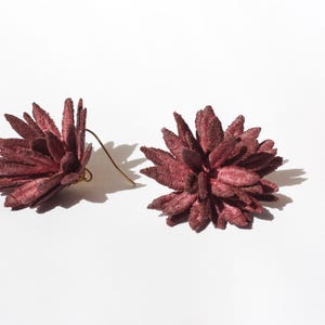 Puede incluir: Un par de pendientes florales de color burdeos. Cada pendiente presenta un dise&ntilde;o texturizado y puntiagudo con un gancho dorado. Los pendientes est&aacute;n sobre un fondo blanco, con sombras.