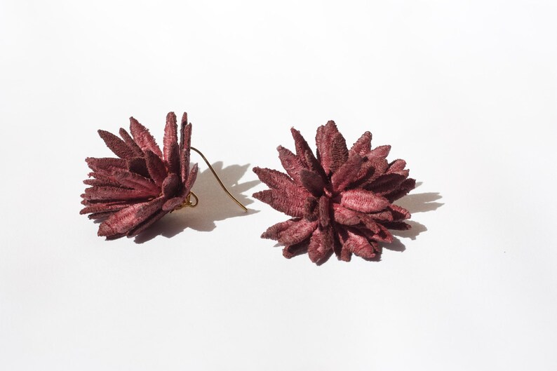 Puede incluir: Un par de pendientes florales de color burdeos. Cada pendiente presenta un dise&ntilde;o texturizado y puntiagudo con un gancho dorado. Los pendientes est&aacute;n sobre un fondo blanco, con sombras.