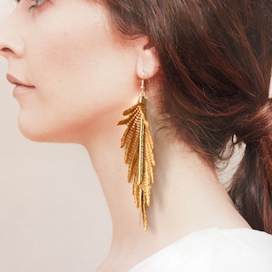 Op de afbeelding: Gouden veren oorbellen met een lang, delicaat ontwerp. De oorbellen hangen en hebben een gouden metalen haak.