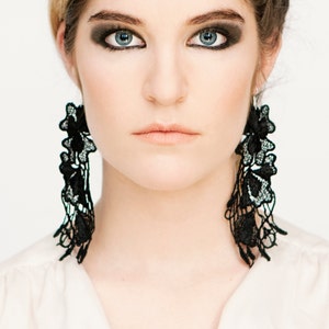 May include: A woman with dark eye makeup is wearing long black lace earrings. The earrings are delicate and intricate, with a floral design.