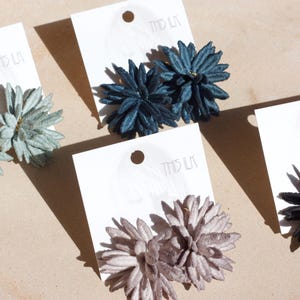 Puede incluir: Primer plano de cuatro pares de pendientes con forma de flor en colores verde claro, azul oscuro, malva y negro. Cada par est&aacute; sujeto a una tarjeta blanca. Los pendientes se muestran sobre una superficie marr&oacute;n claro.