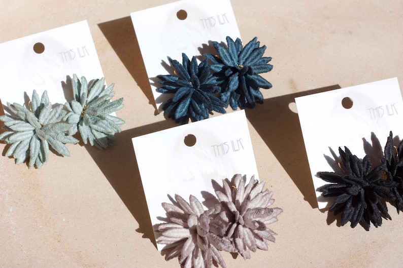 Puede incluir: Primer plano de cuatro pares de pendientes con forma de flor en colores verde claro, azul oscuro, malva y negro. Cada par est&aacute; sujeto a una tarjeta blanca. Los pendientes se muestran sobre una superficie marr&oacute;n claro.