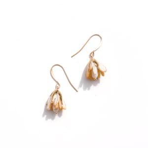 May include: A pair of gold-coloured drop earrings. Each earring features a small, bell-shaped flower with white and gold petals and a small, round pearl at the centre. The earrings are set against a white background.