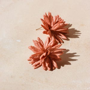 Puede incluir: Dos pendientes de bot&oacute;n florales de color coral. Los pendientes est&aacute;n hechos de un material texturizado, posiblemente tela o bordado, y tienen un dise&ntilde;o espinoso y en capas. Se colocan sobre un fondo neutro y claro.