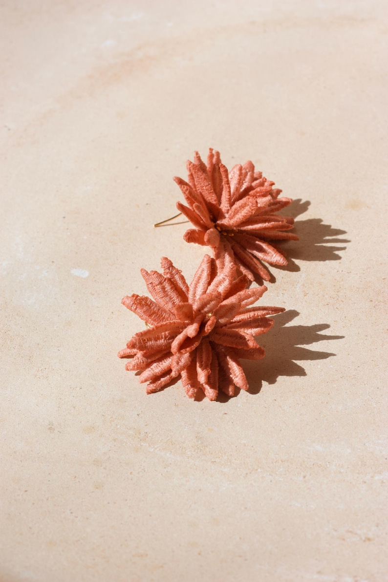 Puede incluir: Dos pendientes de bot&oacute;n florales de color coral. Los pendientes est&aacute;n hechos de un material texturizado, posiblemente tela o bordado, y tienen un dise&ntilde;o espinoso y en capas. Se colocan sobre un fondo neutro y claro.