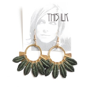 Puede incluir: Un par de pendientes de aro dorados con flecos de tela verde. Los pendientes están en una tarjeta blanca con el texto "THIS ILK" impreso en ella.