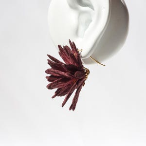 Puede incluir: Un solo pendiente floral burdeos con un gancho dorado. El pendiente est&aacute; sujeto a una pantalla blanca con forma de oreja. El dise&ntilde;o floral tiene un aspecto texturizado y en capas, con m&uacute;ltiples elementos en forma de p&eacute;talos que irradian hacia afuera.