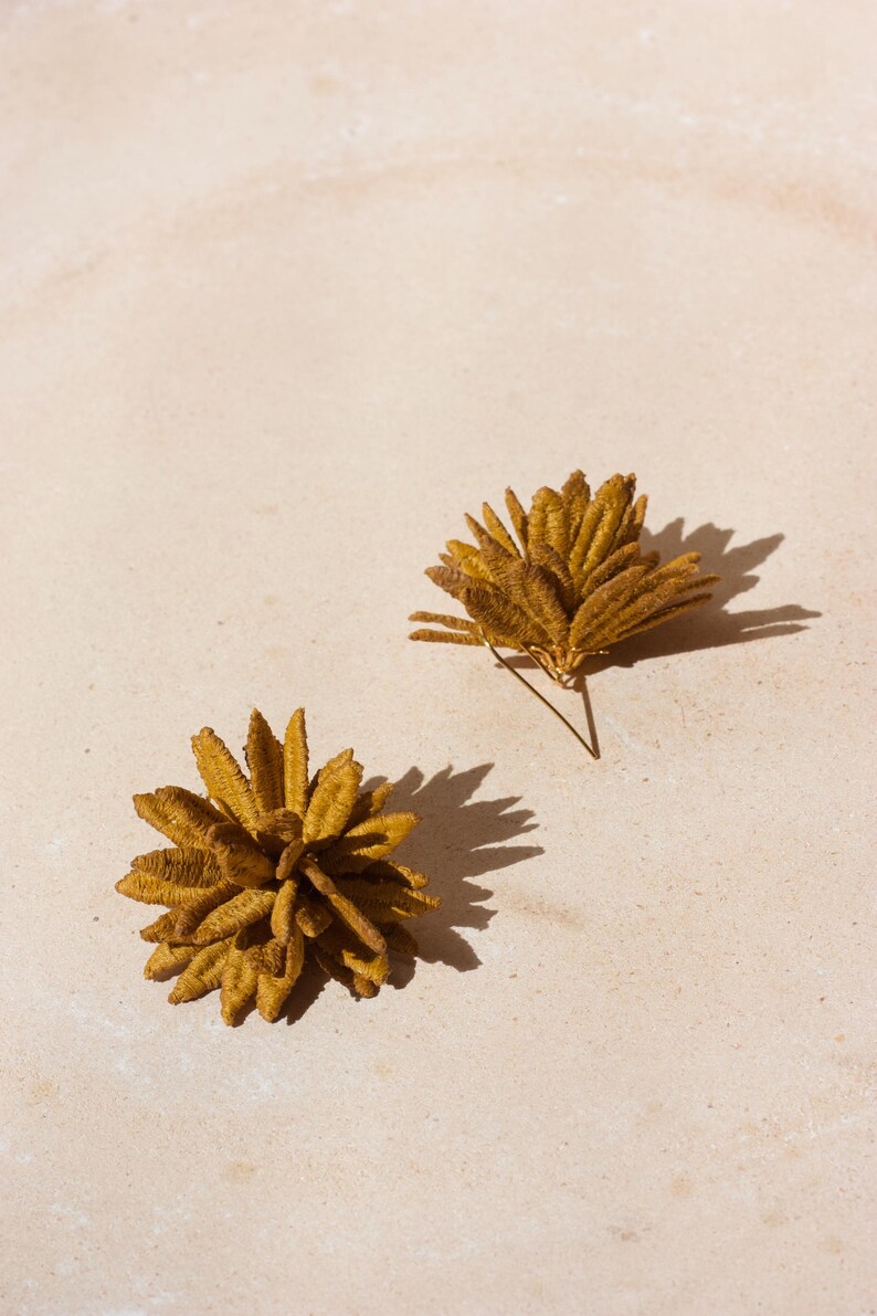 Puede incluir: Dos pendientes dorados con forma de flor. Uno es un pendiente de bot&oacute;n y el otro es un grupo de p&eacute;talos. Los pendientes est&aacute;n sobre un fondo neutro y claro, proyectando sombras.