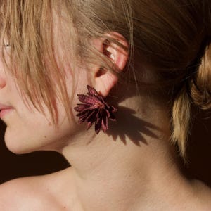 Puede incluir: Primer plano de una oreja adornada con un pendiente en forma de flor, de color burdeos oscuro. El pendiente proyecta una sombra en el cuello de la persona. La persona tiene el pelo claro.