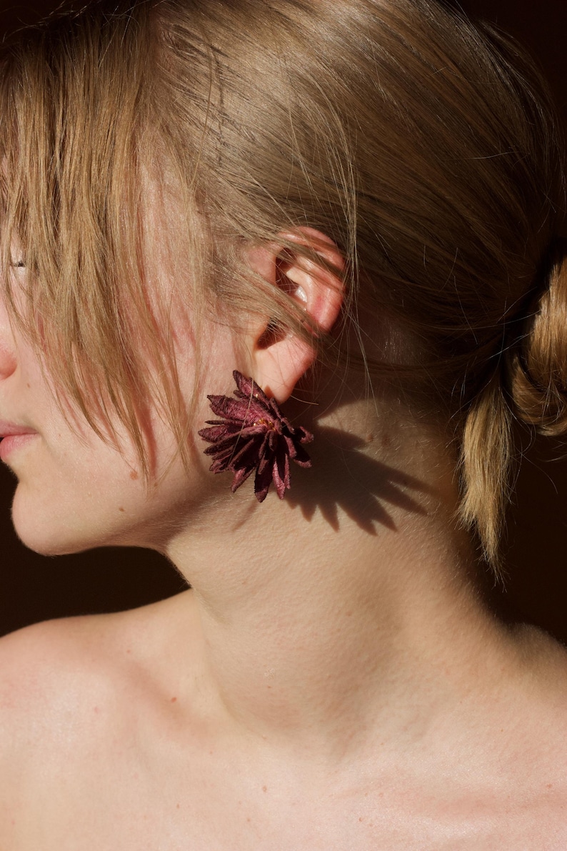 Puede incluir: Primer plano de una oreja adornada con un pendiente en forma de flor, de color burdeos oscuro. El pendiente proyecta una sombra en el cuello de la persona. La persona tiene el pelo claro.