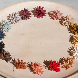 Puede incluir: Una colecci&oacute;n de pendientes de flores hechos a mano, dispuestos en semic&iacute;rculo sobre un plato de cer&aacute;mica beige. Los pendientes son de varios colores, incluyendo rojo, blanco y azul. Los pendientes son un art&iacute;culo artesanal.