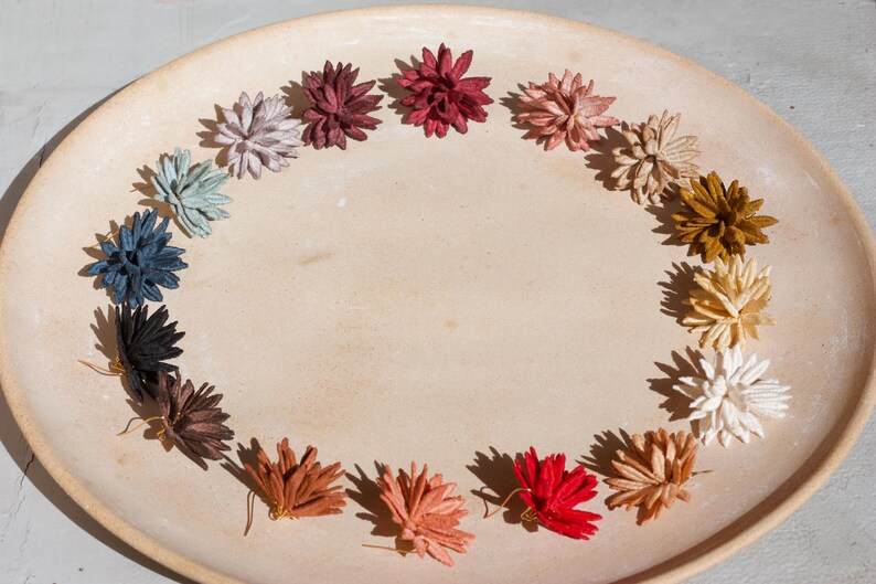 Puede incluir: Una colecci&oacute;n de pendientes de flores hechos a mano, dispuestos en semic&iacute;rculo sobre un plato de cer&aacute;mica beige. Los pendientes son de varios colores, incluyendo rojo, blanco y azul. Los pendientes son un art&iacute;culo artesanal.