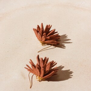 Puede incluir: Dos pendientes puntiagudos de color terracota con alambres dorados se muestran sobre una superficie beige claro. Los pendientes proyectan sombras, lo que sugiere la luz del sol. Los pendientes tienen un dise&ntilde;o floral o de estrella.