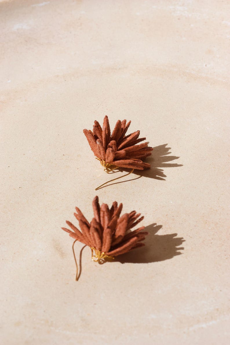 Puede incluir: Dos pendientes puntiagudos de color terracota con alambres dorados se muestran sobre una superficie beige claro. Los pendientes proyectan sombras, lo que sugiere la luz del sol. Los pendientes tienen un dise&ntilde;o floral o de estrella.