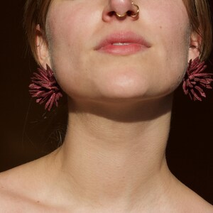 Puede incluir: Primer plano de pendientes espinosos de color burdeos. Los pendientes tienen un dise&ntilde;o texturizado y en capas, que se asemeja a una flor o una explosi&oacute;n. La imagen se centra en los pendientes y la parte inferior de la cara y el cuello de una persona.
