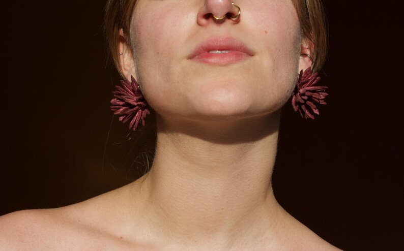 Puede incluir: Primer plano de pendientes espinosos de color burdeos. Los pendientes tienen un dise&ntilde;o texturizado y en capas, que se asemeja a una flor o una explosi&oacute;n. La imagen se centra en los pendientes y la parte inferior de la cara y el cuello de una persona.