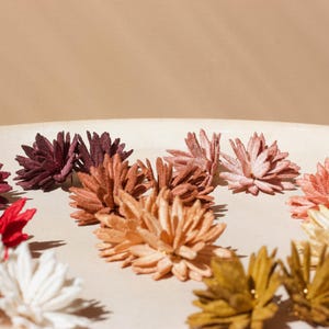Puede incluir: Una colecci&oacute;n de decoraciones florales coloridas y hechas a mano en tonos de rojo, blanco, burdeos, melocot&oacute;n y dorado. Las flores est&aacute;n dispuestas sobre una superficie redonda de color beige claro, creando una exhibici&oacute;n visualmente atractiva.
