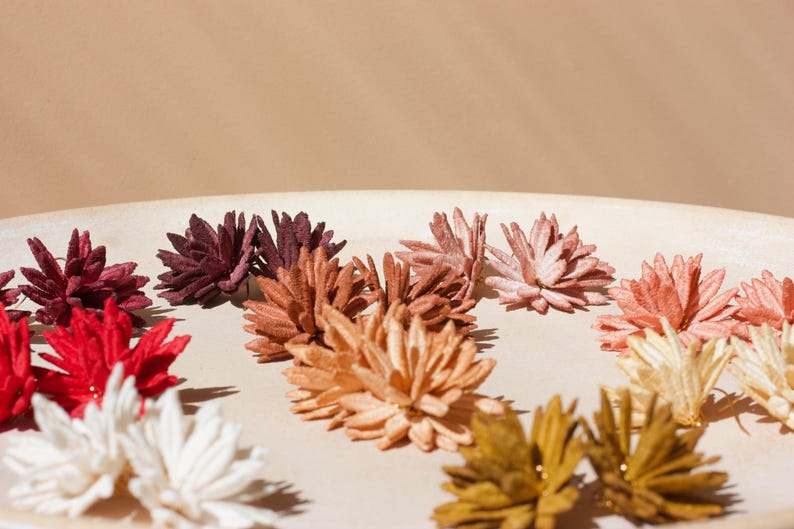Puede incluir: Una colecci&oacute;n de decoraciones florales coloridas y hechas a mano en tonos de rojo, blanco, burdeos, melocot&oacute;n y dorado. Las flores est&aacute;n dispuestas sobre una superficie redonda de color beige claro, creando una exhibici&oacute;n visualmente atractiva.