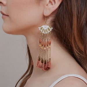 May include: A pair of long, dangling earrings with a white crocheted fan-shaped top, brown wooden beads, and pink tassels.