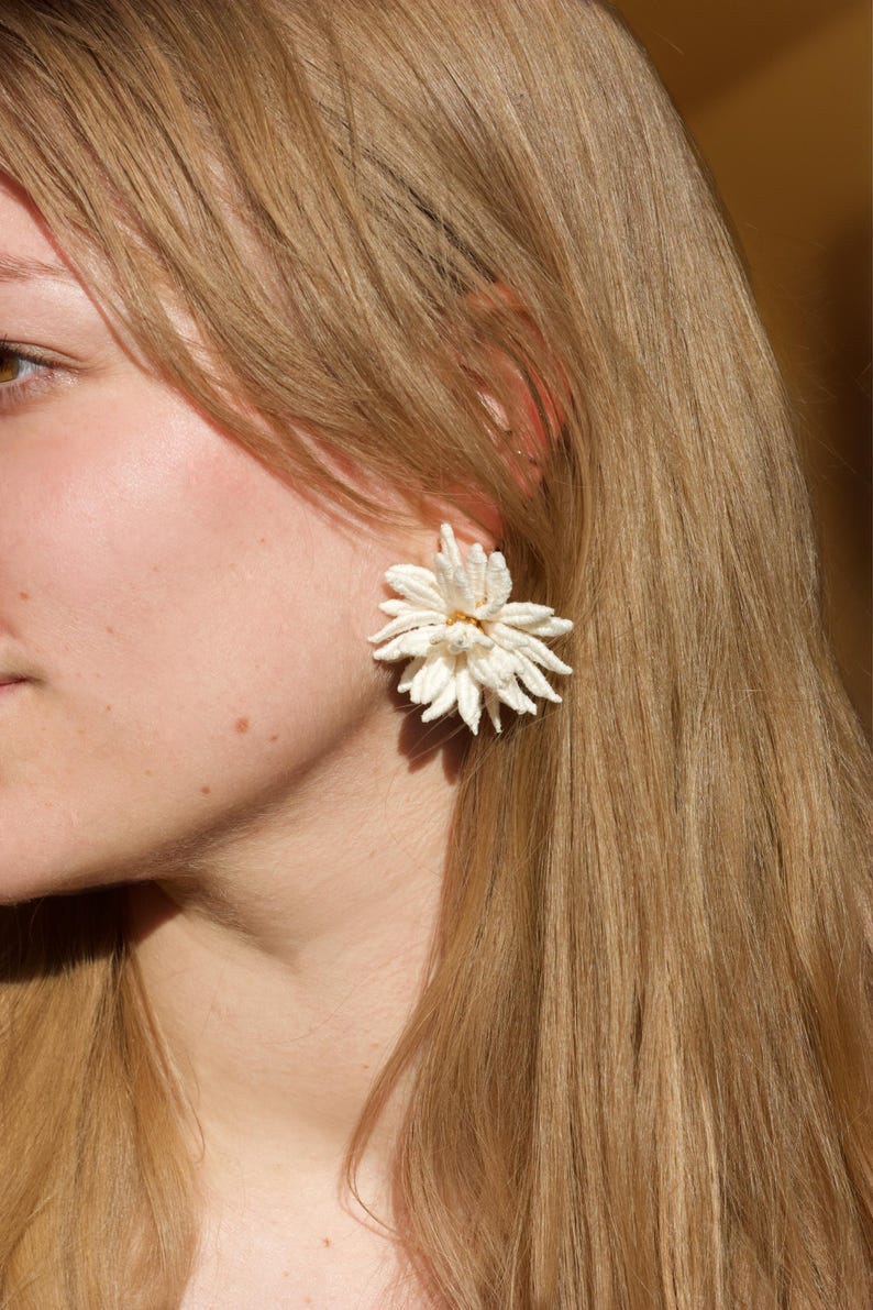 Puede incluir: Un pendiente blanco con forma de flor y superficie texturizada. El pendiente se lleva en la oreja y tiene un peque&ntilde;o centro dorado. Un accesorio &uacute;nico.