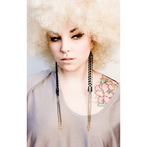 Op de afbeelding: Een vrouw met een blond afro-pruik draagt een grijs T-shirt en zwarte en gouden kwast-oorbellen. Ze heeft een klein neusringetje en een roze bloemtattoo op haar schouder.