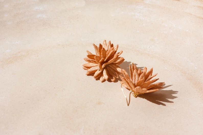 Puede incluir: Un par de pendientes artesanales en forma de flor, de color marr&oacute;n claro, con ganchos dorados. Los pendientes est&aacute;n hechos de un material texturizado y se encuentran sobre un fondo claro y neutro.