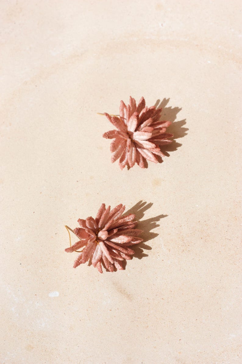 Puede incluir: Dos pendientes en forma de flor espinosa, de color rosa empolvado. Los pendientes est&aacute;n sobre una superficie beige claro, proyectando sombras. Tienen un aspecto delicado y texturizado, lo que sugiere un material de tela o papel.