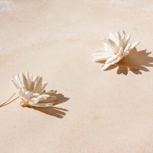 Puede incluir: Dos pendientes blancos con forma de flor y postes dorados se muestran sobre una superficie beige claro. Los pendientes proyectan sombras oscuras y la imagen est&aacute; iluminada con luz natural, creando una est&eacute;tica suave y neutra.