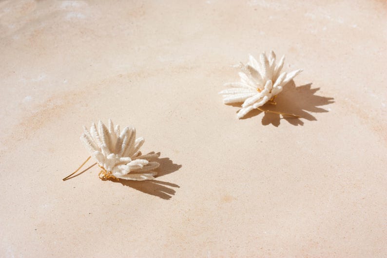 Puede incluir: Dos pendientes blancos con forma de flor y postes dorados se muestran sobre una superficie beige claro. Los pendientes proyectan sombras oscuras y la imagen est&aacute; iluminada con luz natural, creando una est&eacute;tica suave y neutra.