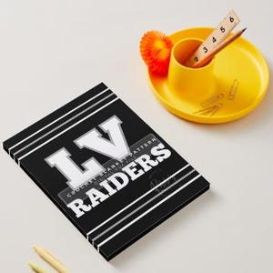May include: A black rectangular document featuring white horizontal stripes and the bold white text LV RAIDERS, with a smaller label reading CROCHET BLANKET PATTERN DIGITAL PDF FILE. A wooden ruler shows a length of 6 inches near a yellow desk organizer.