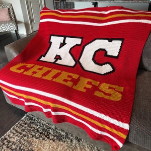 May include: A red crochet blanket featuring the white letters KC with a black outline and the word CHIEFS in yellow lettering. The blanket has horizontal stripes in white and yellow at the top and bottom edges, creating a sports-themed design.