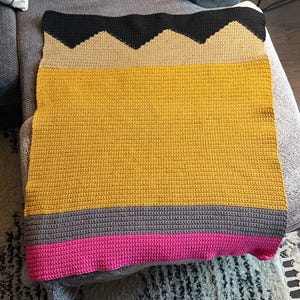 Può includere: Una coperta rettangolare all'uncinetto progettata per assomigliare a una matita. La parte superiore presenta un motivo a zigzag nero, seguito da una sezione color marrone chiaro, un grande corpo giallo, una fascia grigia e un bordo rosa.