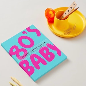 May include: A rectangular document with a bright turquoise cover featuring the large hot pink text 80's BABY and smaller black text CROCHET THROW DESIGN DIGITAL PATTERN ONLY. Nearby is a yellow circular tray with a wooden ruler and pencil.