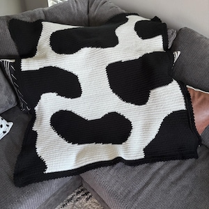 May include: A rectangular crochet blanket featuring a high-contrast cow print pattern. The design consists of large, irregular black patches set against a solid white background, created with a consistent, textured stitch throughout the fabric.