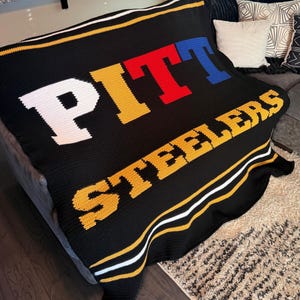 Może przedstawiać: Czarny szydełkowany koc z napisem PITT wykonanym z białych, żółtych, czerwonych i niebieskich blokowych liter oraz napisem STEELERS z żółtych liter poniżej. Koc posiada poziome białe i żółte paski wzdłuż górnej i dolnej krawędzi.